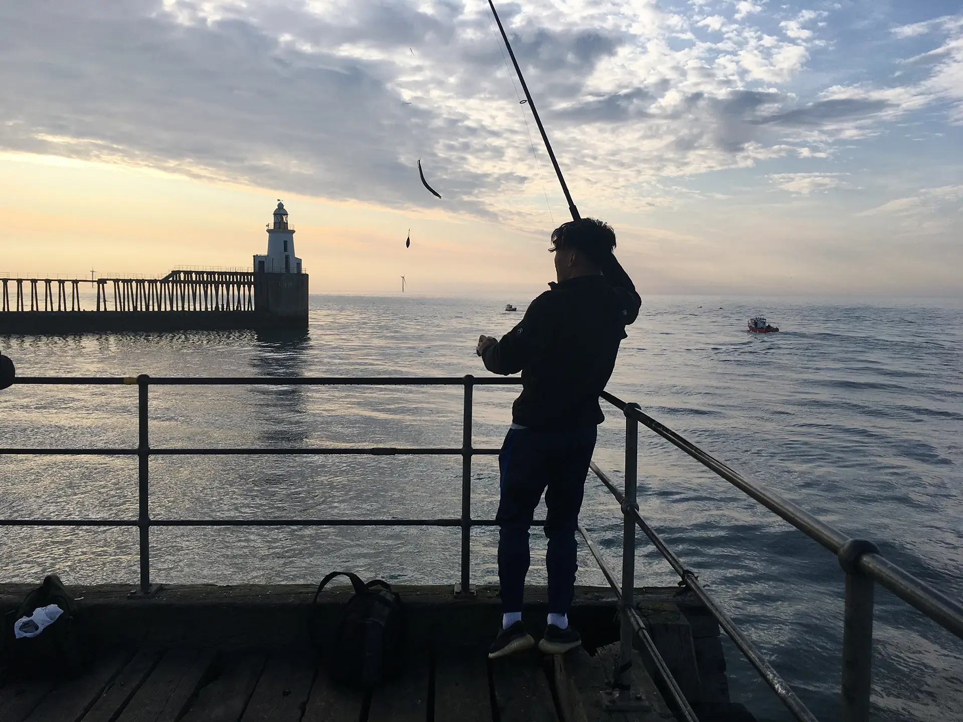 Angler catching a fish silhouetted on an evening with pier and lighthouse in the background.