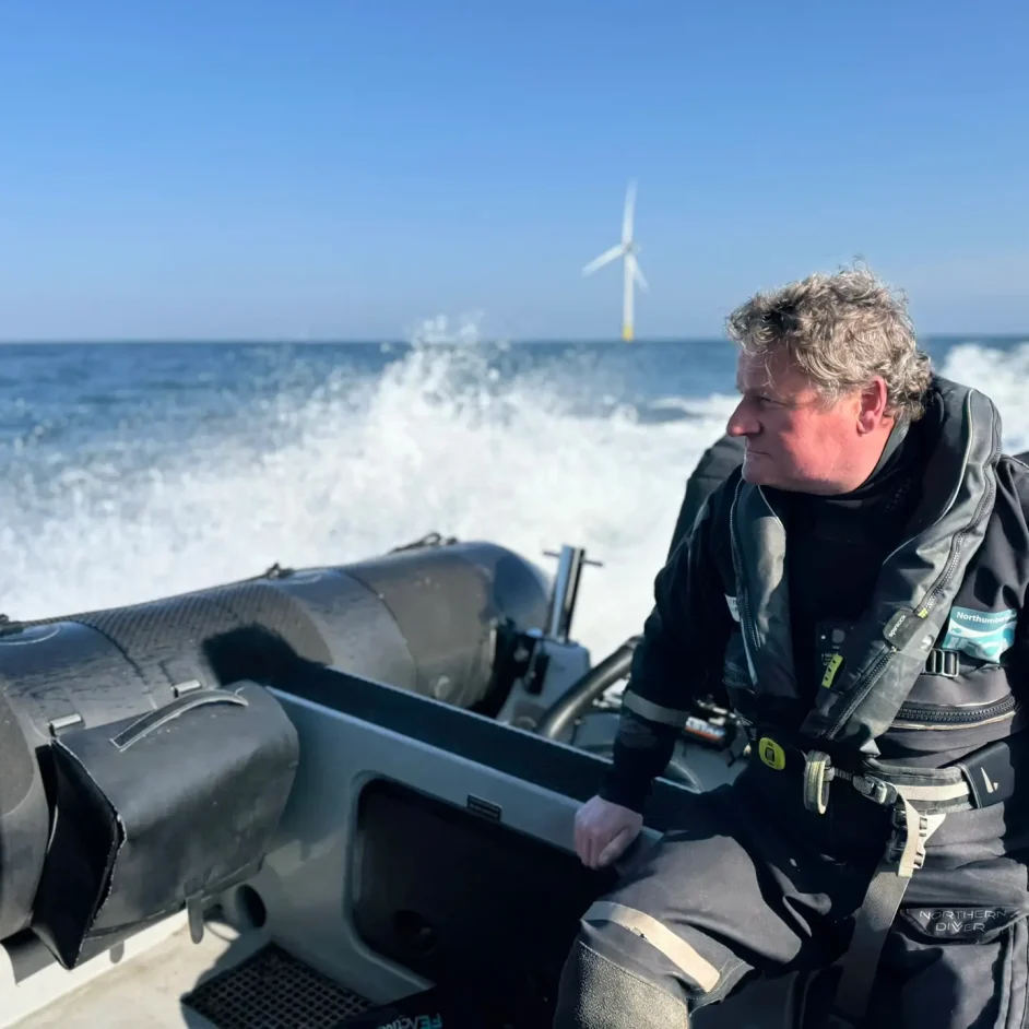 Man wearing NIFCA branded workwear at sea on patrol vessel with blue sea and wind turbines in background.