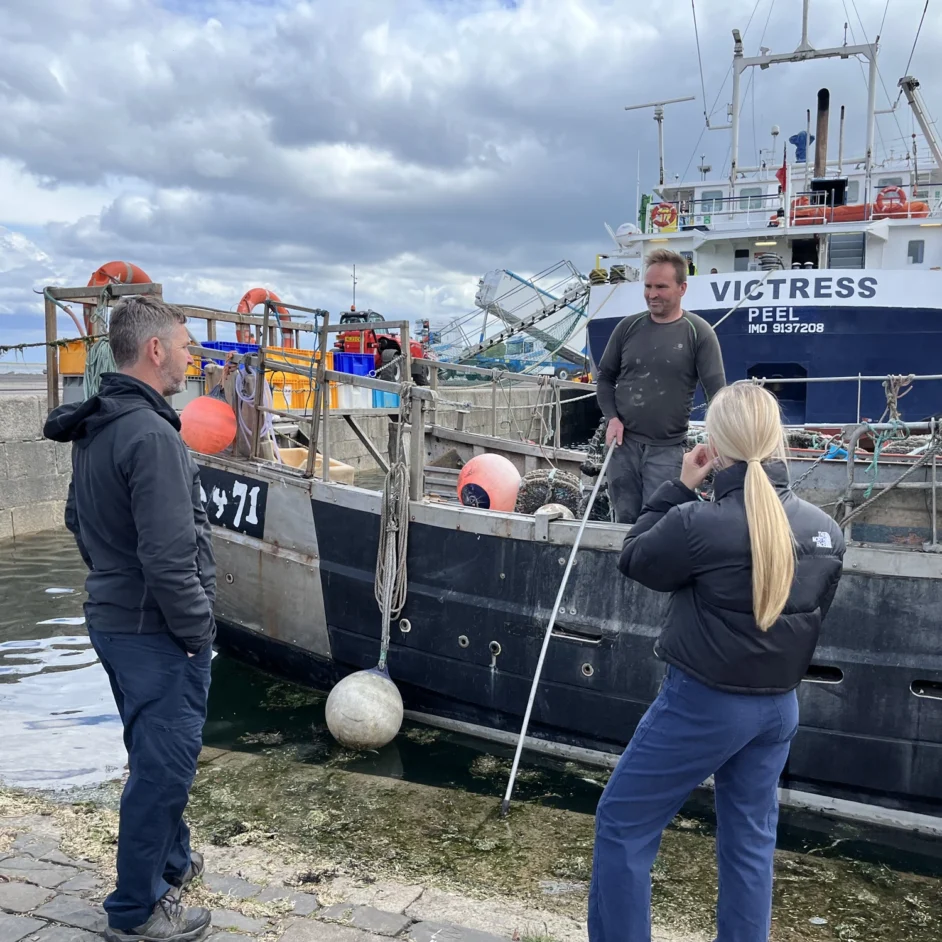 Two NIFCA representatives talk to fisher who is stood on a boat at a dock in Berwick.