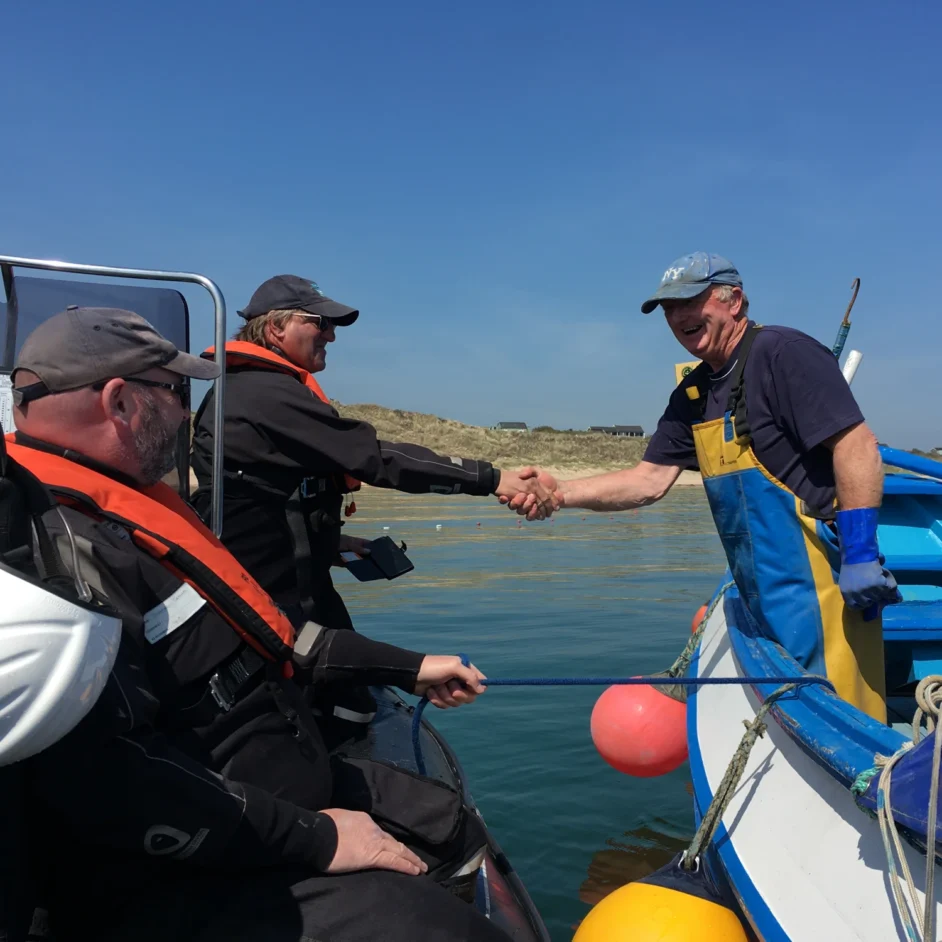 Two NIFCA officers on patrol boat shaking hands with fisher on another boat.