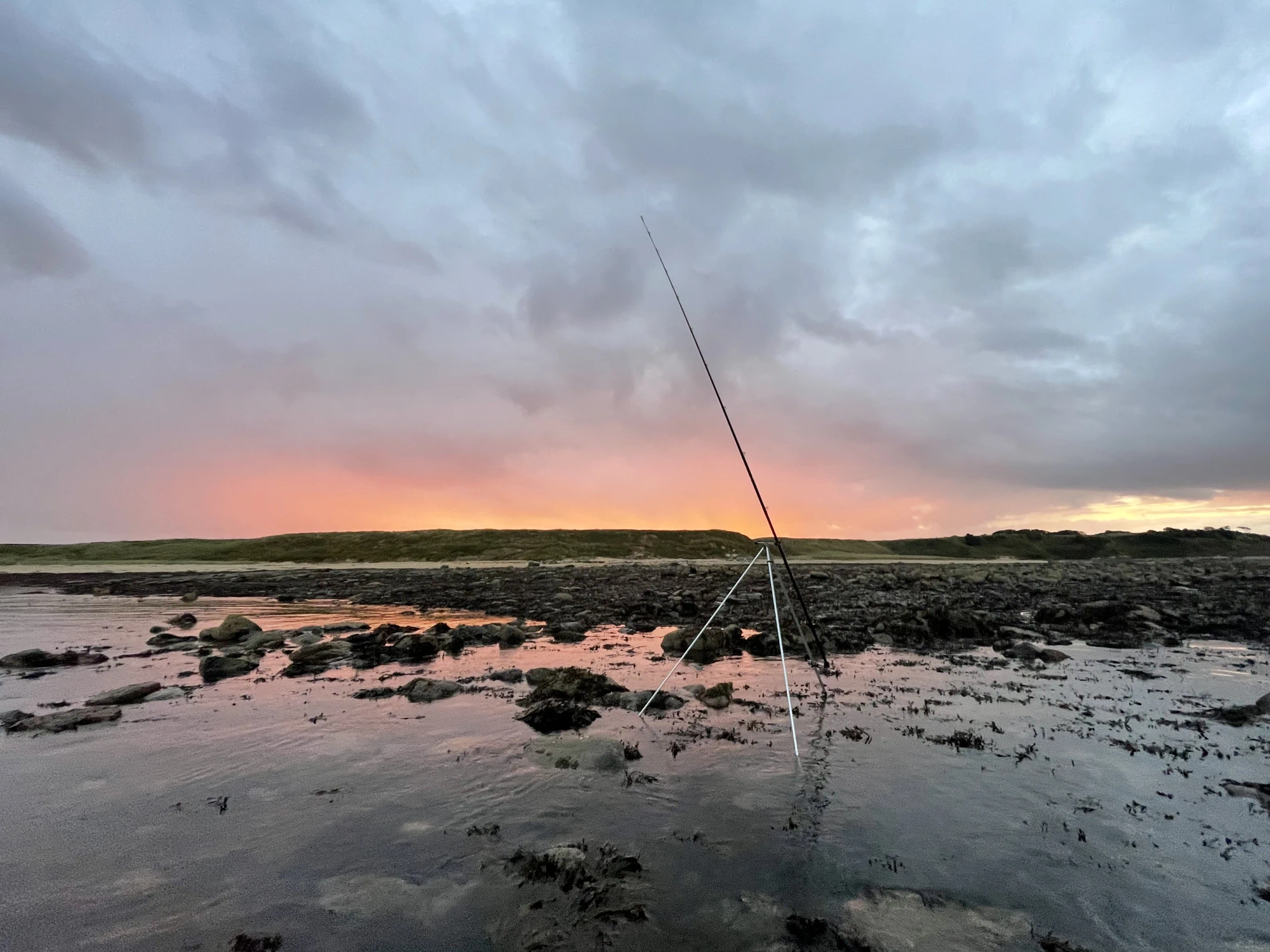 Beautiful sunset in Northumberland with pink sky reflected on water and fishing rod set up on rocky shore.