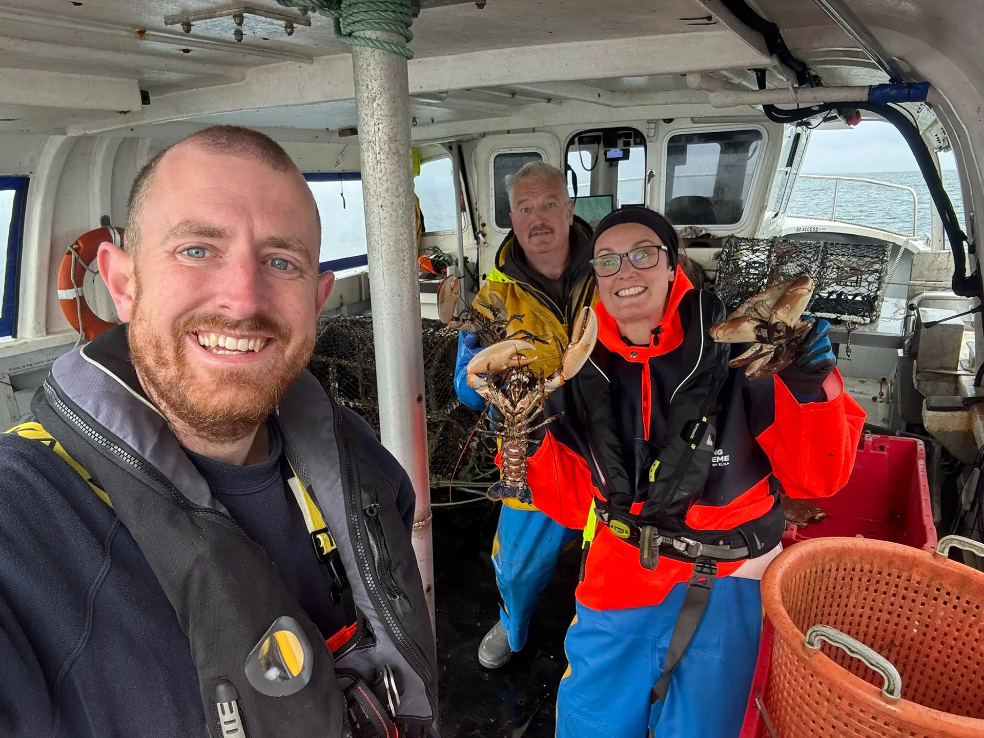 Three NIFCA crew members smiling at camera holding lobsters in vessel cabin during a survey.